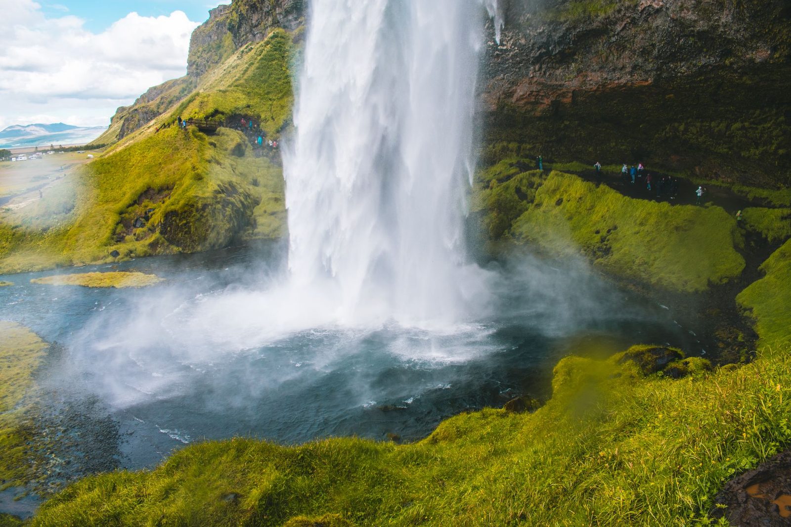 A misty Seljalandsfoss waterfall with a rainbow visible through the mist, surrounded by lush greenery